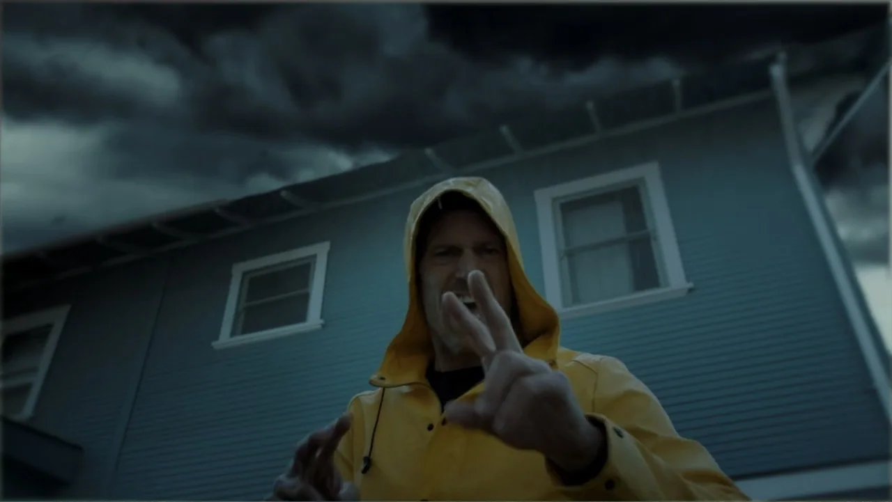 Storm Damage (2000) movie still: medium — A man stands in front of a blue-sided house under a heavy, dark sky. The man is wearing a bright yellow…