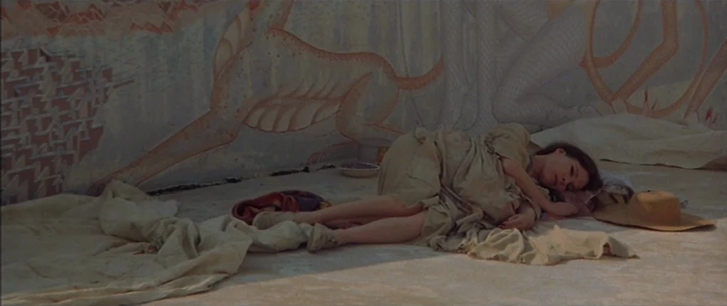 3 Women (1977) movie still: wide — A woman lies on a concrete floor in front of a painted mural. She rests on fabric scraps with a straw hat placed…