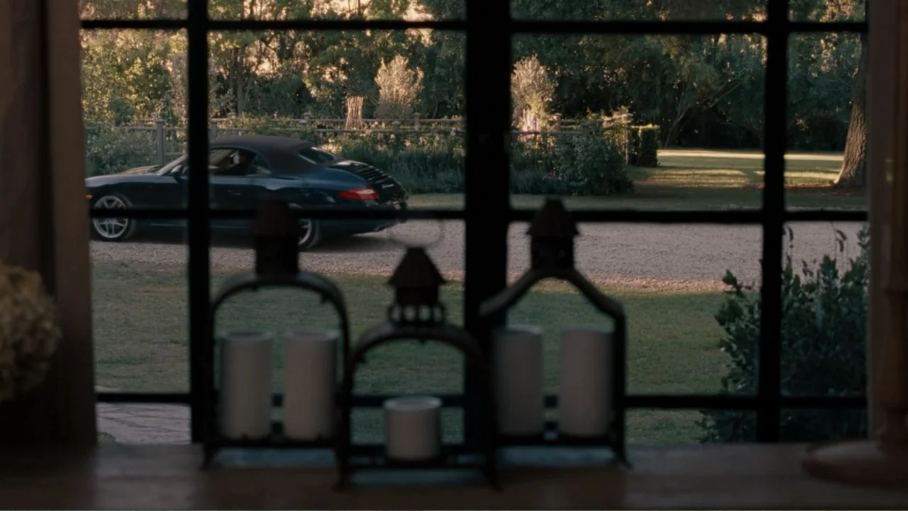It's Complicated (2009) movie still: medium — A view through a window grid reveals a gravel driveway and green trees. A dark convertible car passes…