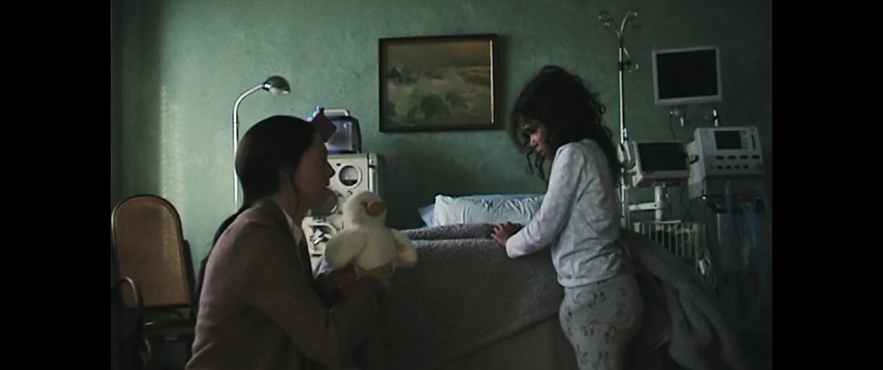Birth/Rebirth (2023) movie still: medium — A dimly lit room with muted green walls contains medical equipment and a bed. An adult woman approaches a…