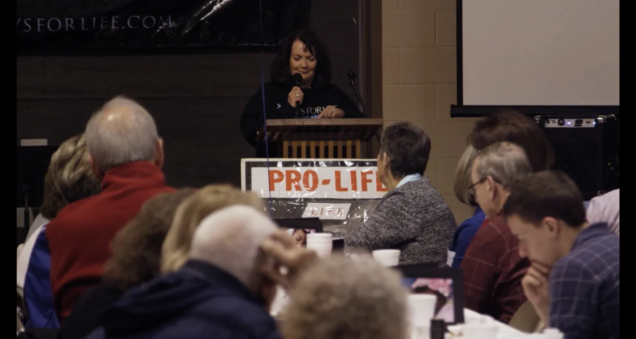 Abortion: Stories Women Tell (2016) movie still: medium — A woman stands behind a wooden podium inside a room with a projector screen. The audience…