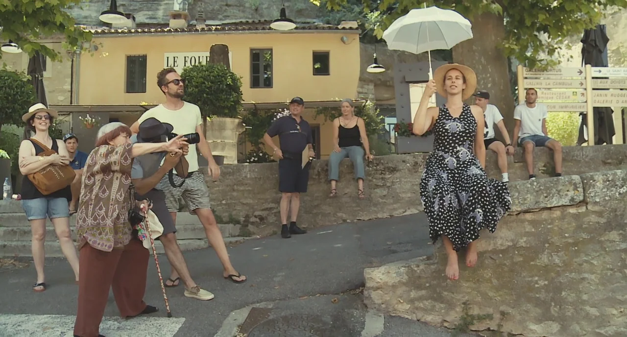 Faces Places (2017) movie still: wide — A stone-walled outdoor plaza in a European village features a yellow stucco building. A woman sits on a low…