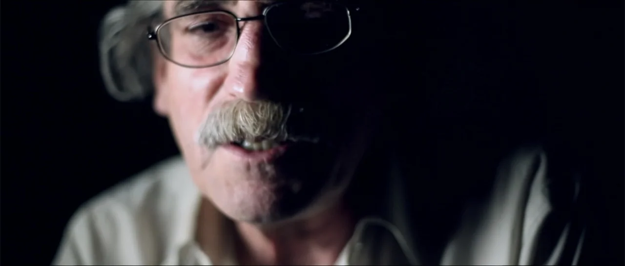 Garnet’s Gold (2014) movie still: closeup — A dimly lit interior space with the subject centered against a black void. An older man with a mustache…