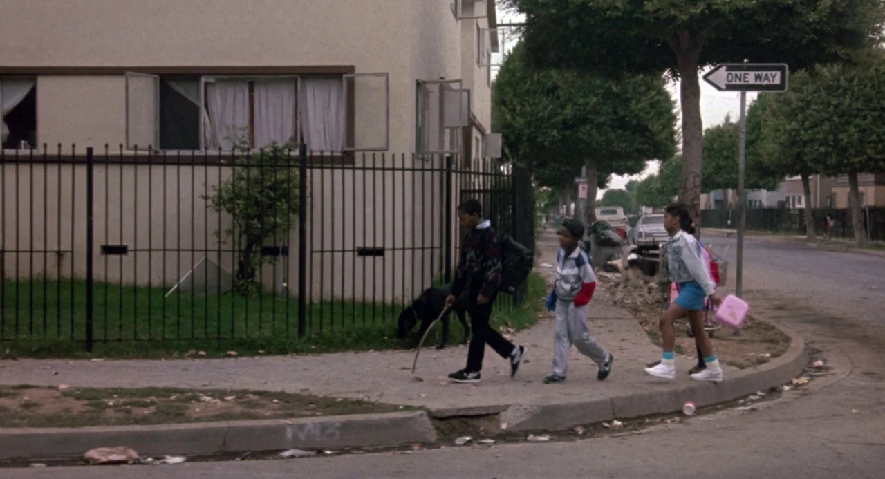 Boyz In The Hood (1991) movie still: wide — A residential sidewalk passes by a light-colored stucco apartment building with a black fence. Three…