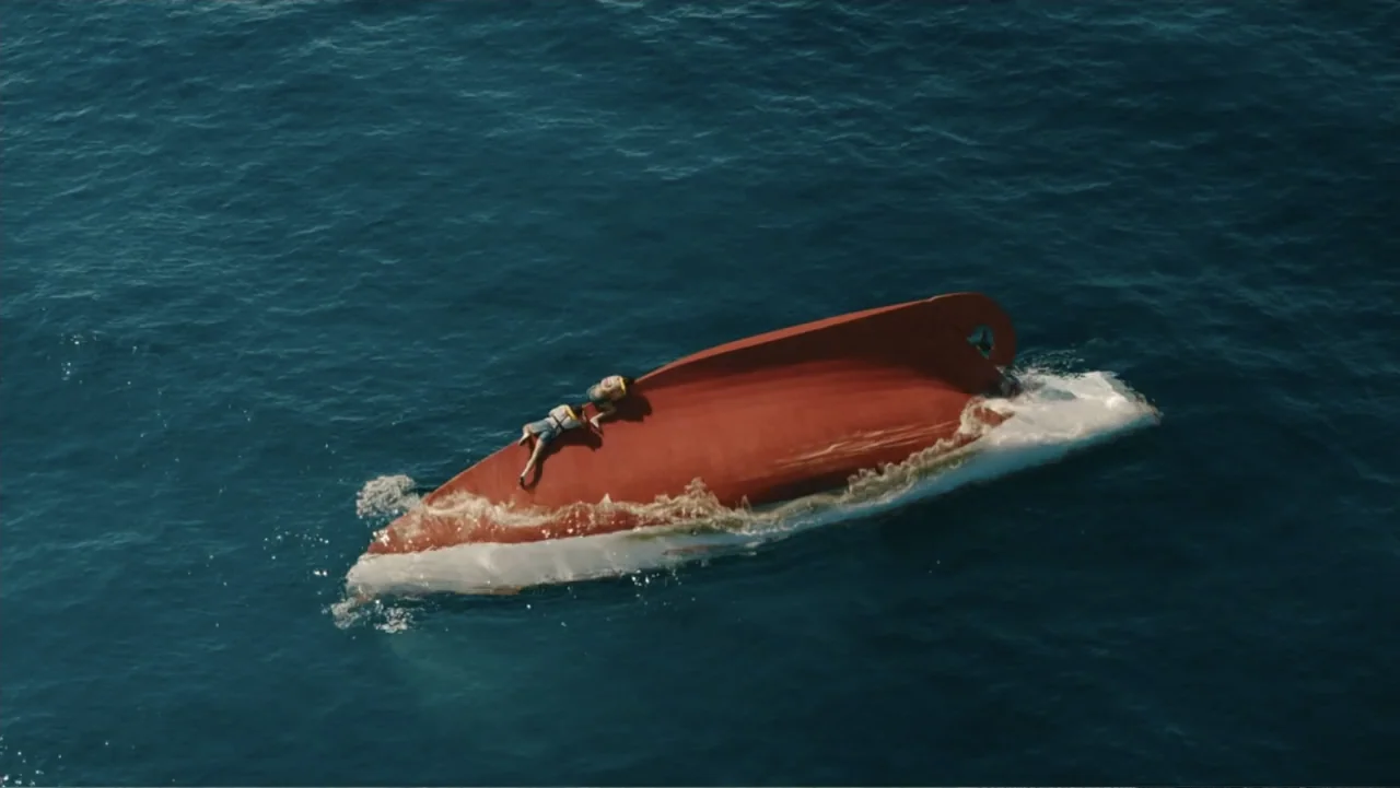 Jurassic World Rebirth (2025) movie still: wide — An overturned boat hull drifts through calm dark blue water in open ocean. Two individuals in life…