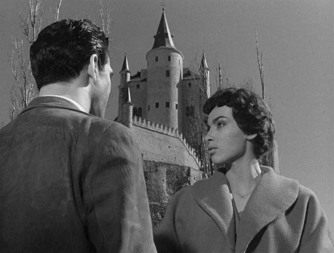 Mr. Arkadin (1955) movie still: medium — A stone castle with pointed towers stands behind two people standing in a field. The couple faces each…