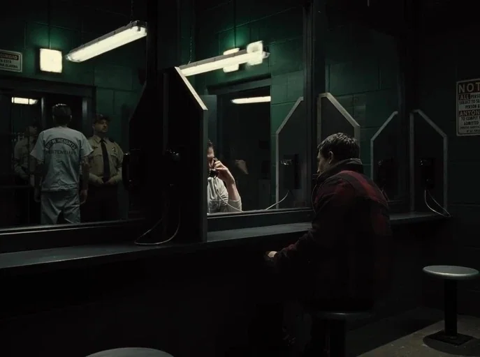 Zack Snyder's Justice League (2021) movie still: medium — A dark visitation room with cinder block walls and glass dividers. A man sits at a small…