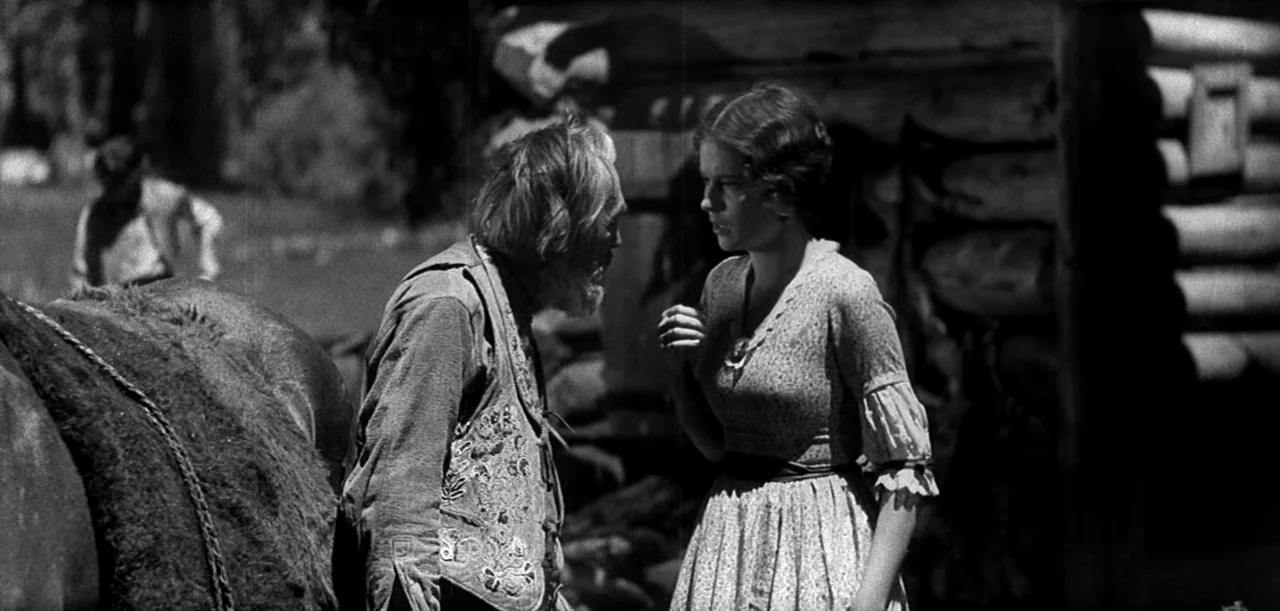 The Big Trail (1930) movie still: medium — An outdoor area with a rustic log cabin structure visible in the background. An elderly man in a vest…