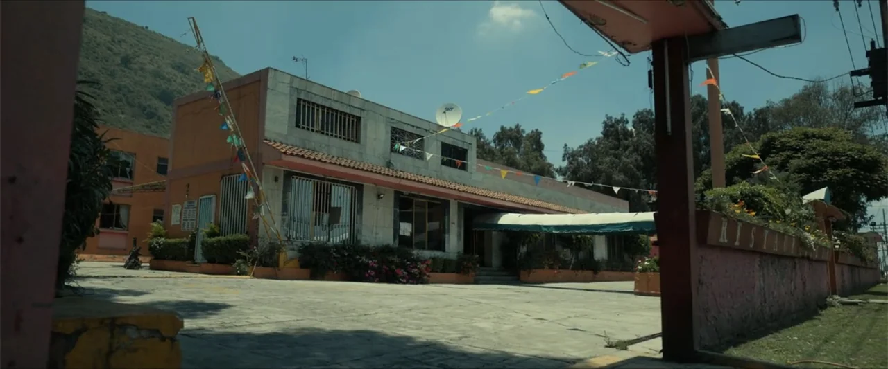 Dive (2013) movie still: establishing — A low-slung, two-story commercial building with light stone walls stands near a mountain. Decorative…