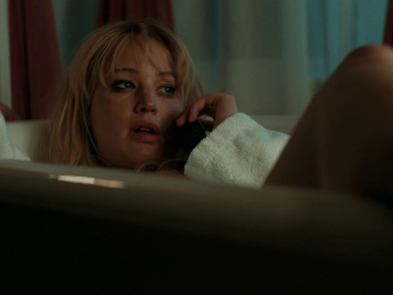 Die My Love (2025) movie still: closeup — A woman rests in a dimly lit bathroom while partially submerged in a tub. She holds a small dark mobile…
