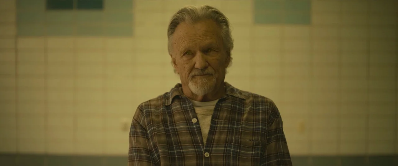 Blaze (2018) movie still: medium — An older man stands before a wall covered in pale green square tiles. He stares forward with an unblinking,…