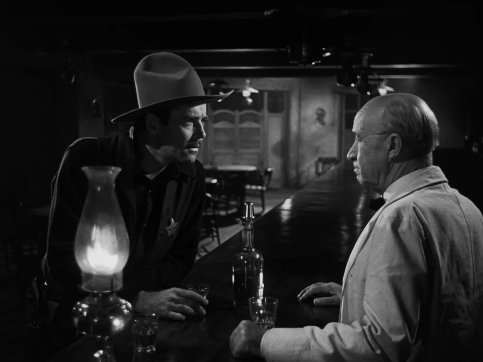My Darling Clementine (1946) movie still: medium — A dark wooden bar counter contains a glass bottle and a lit oil lamp. A lawman in a hat faces an…