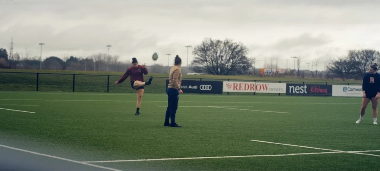 No Woman No Try (2022) movie still: wide — A green synthetic sports field under an overcast grey sky with perimeter fencing. Three women in athletic…