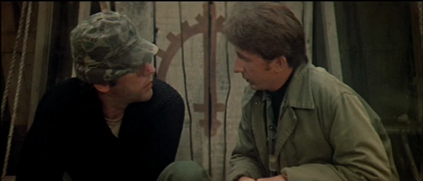 MASH (1970) movie still: medium — Two men sit close together within a wooden, enclosed structure with vertical slats. One man wears a camouflage cap…