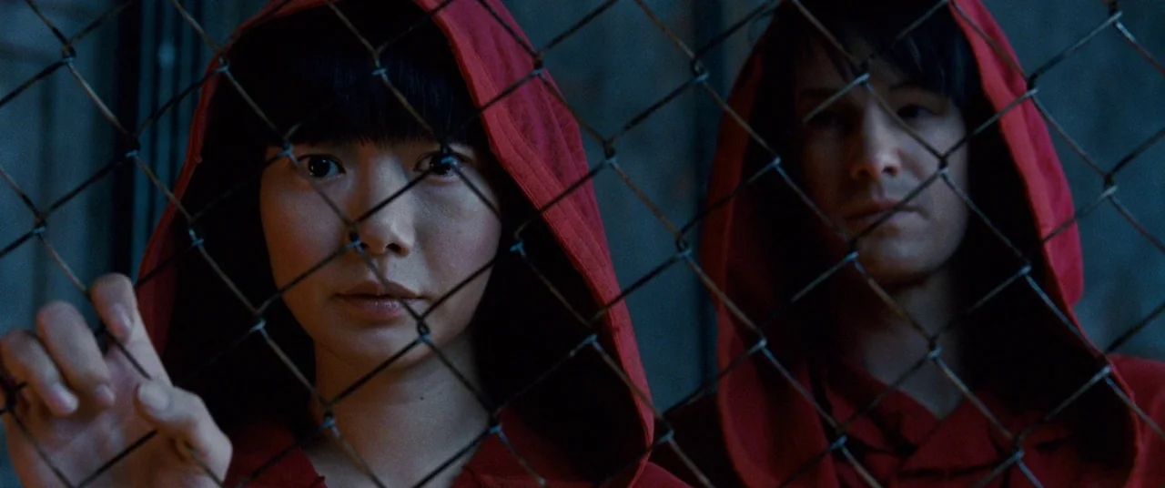 Cloud Atlas (2012) movie still: medium — A dark, textured chain link fence stands between the camera and two hooded subjects. The subjects stare…