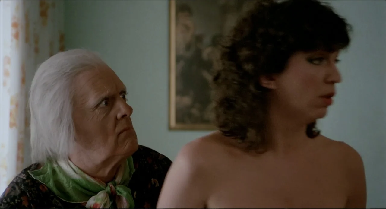 Waiting for the Hearse (1985) movie still: medium — A room with light blue walls featuring a framed painting on the back wall. An elderly woman…