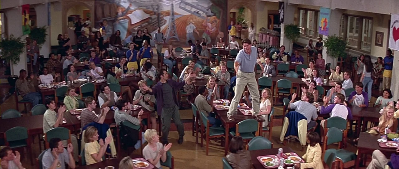 Scream 2 (1997) movie still: wide — A crowded university dining hall features wooden tables, green chairs, and a large wall mural. A young man…