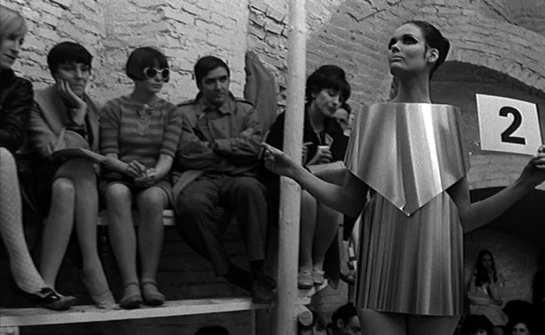 Who Are You, Polly Maggoo? (1966) movie still: medium — A minimalist indoor runway space features rough brick walls and wooden seating for…