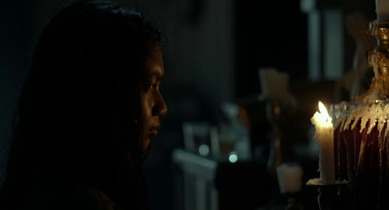 In My Mother’s Skin (2023) movie still: closeup — A dark, dimly lit indoor space with rows of candles in the foreground. A young woman in profile…