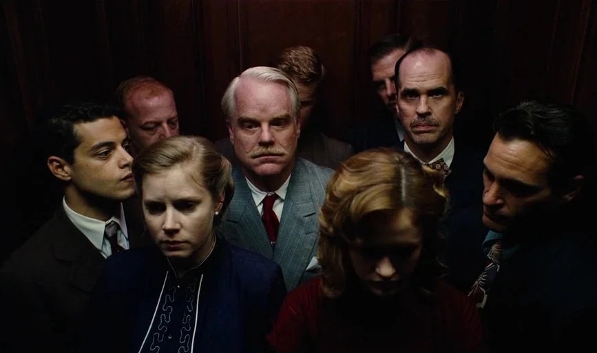 The Master (2012) movie still: medium — A group of people stand closely packed inside a wood-paneled interior space. They face forward in silence…