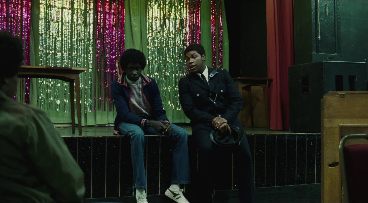 Red, White & Blue (2020) movie still: medium — An indoor community center hall features a small wooden stage with shiny fringe curtains. Two young…