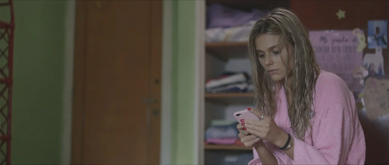Nahir (2024) movie still: medium — A woman sits inside a bedroom with green walls and an open closet behind her. She looks down at a pink smartphone…