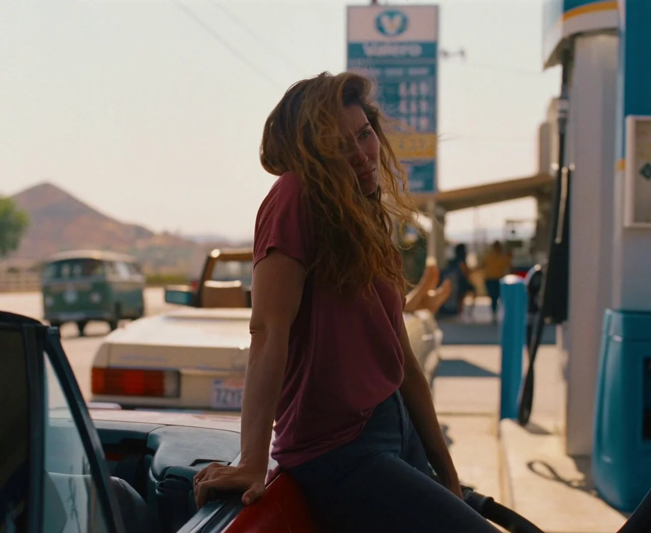 Monica (2022) movie still: medium — A woman leans against a convertible parked at a sunny gas station. Distant hills and vintage cars are visible in…