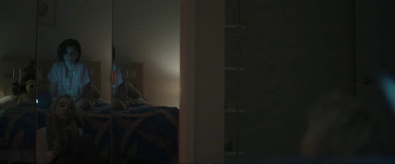 Come True (2020) movie still: medium — A dimly lit room at night containing a bed reflected in a mirror. A young girl sits on the edge of the bed…