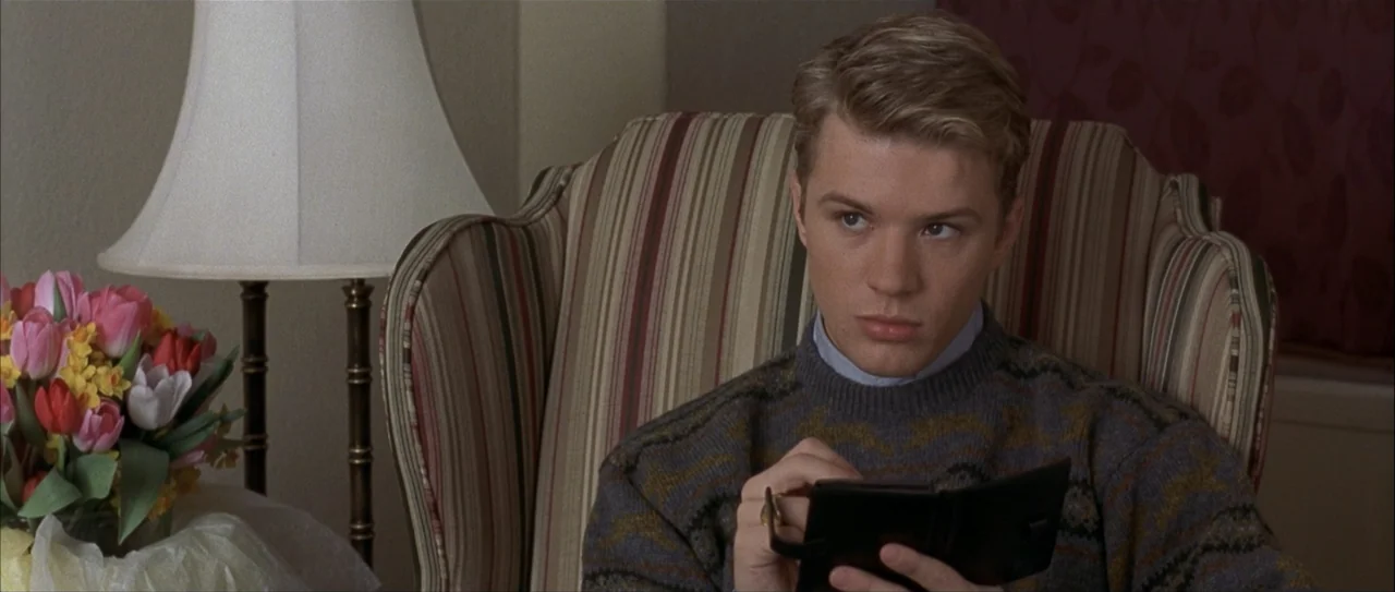 Igby Goes Down (2002) movie still: medium — A living room interior features a striped armchair, a table lamp, and flowers. A young blond man sits in…