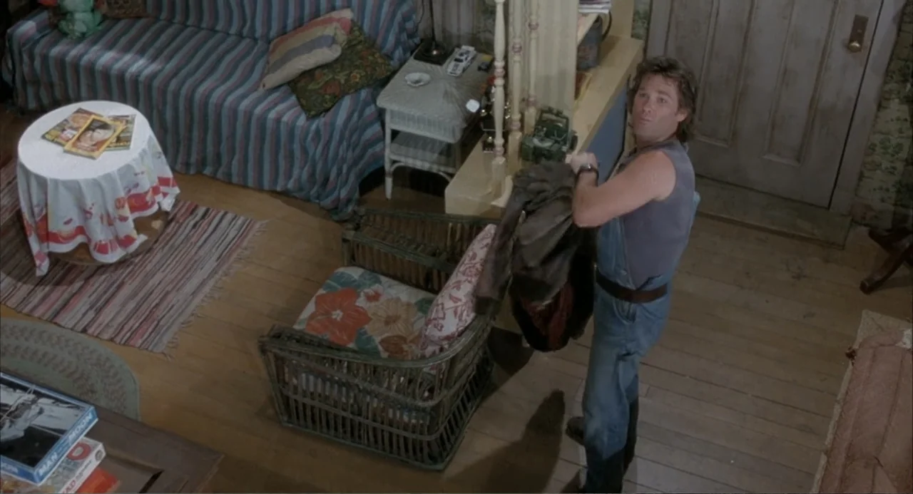 Overboard (1987) movie still: medium — A living room contains a striped couch, a wicker armchair, and a small round table. A man stands near the…