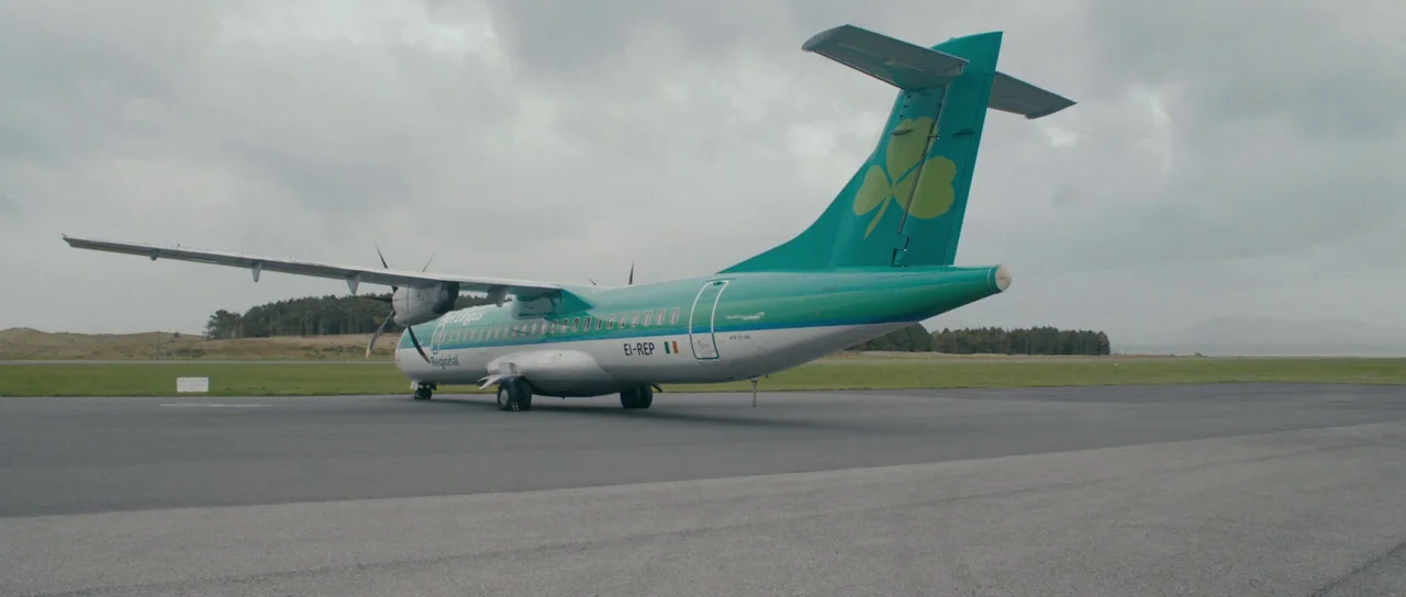 Calvary (2014) movie still: wide — An Aer Lingus Regional airplane rests on an asphalt runway surrounded by grassy fields. The sky is filled with…