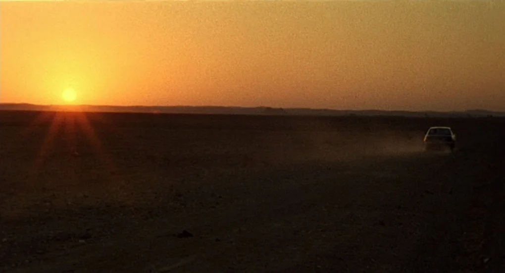 Furia (1999) movie still: wide — A flat desert landscape stretches to the horizon under a bright sunset. A small car drives across the dry ground in…