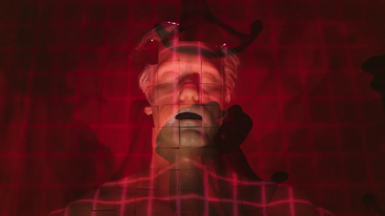 Infinity Pool (2023) movie still: closeup — A dark environment is illuminated by a projected red grid pattern. A single mannequin head occupies the…