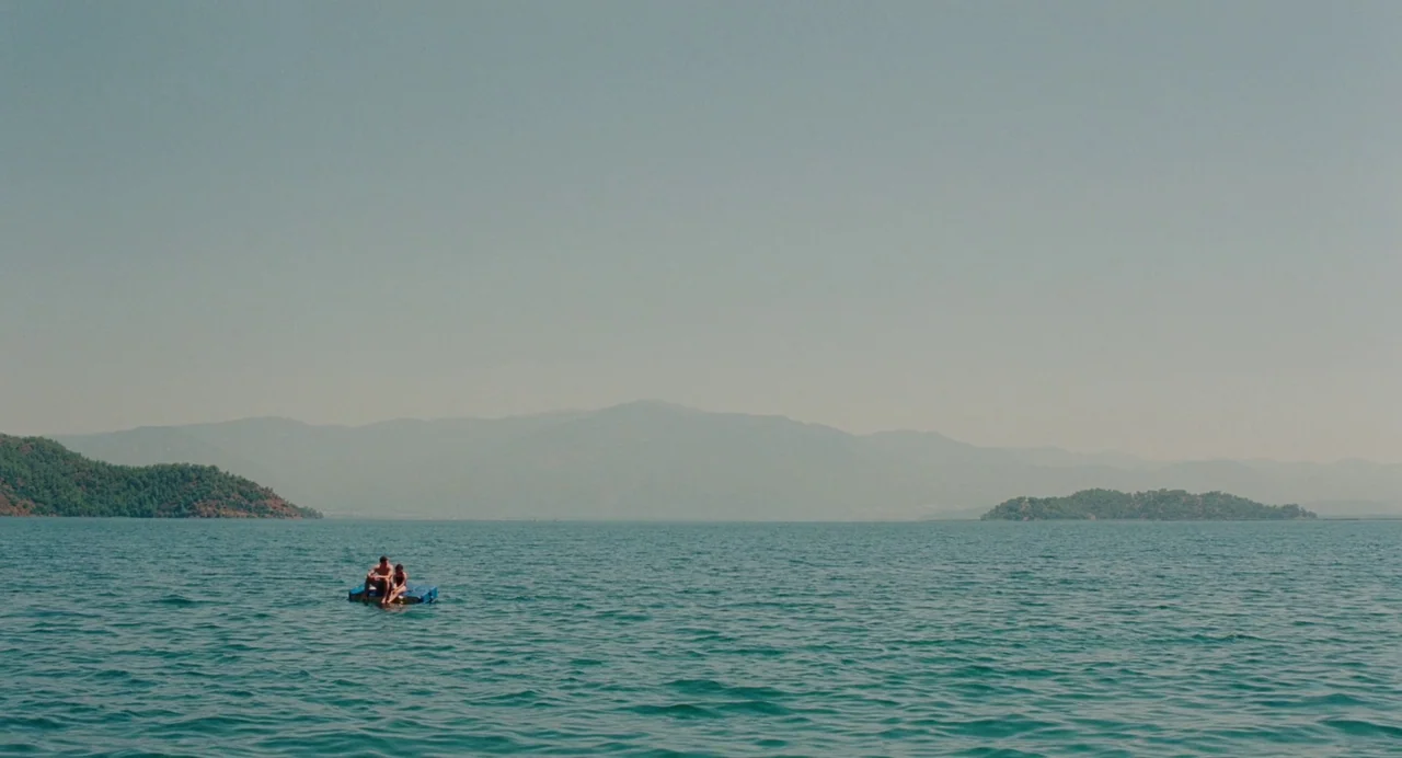 Aftersun (2022) movie still: wide — A vast expanse of water is framed by hazy, distant mountains on the horizon. Two people sit closely together on…