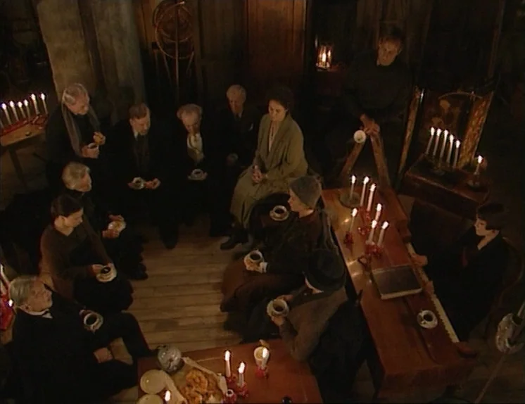 In The Presence of a Clown (1997) movie still: wide — A dimly lit room features a large group of people seated in a circle. They hold mugs and…