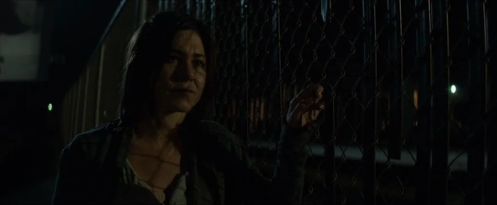 Cake (2014) movie still: medium — A woman stands before a dark chain link fence at night. She grips the metal wires with her fingers while looking…