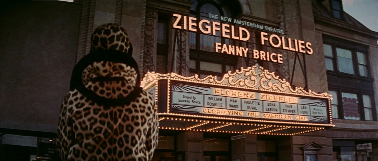 Funny Girl (1968) movie still: medium — The exterior of the New Amsterdam Theatre at night with a bright, illuminated marquee. A person wearing a…