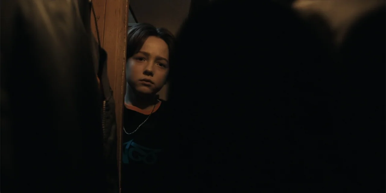 The Monkey (2025) movie still: closeup — A dark, dimly lit indoor space viewed through a slightly ajar wooden door frame. A young person looks…