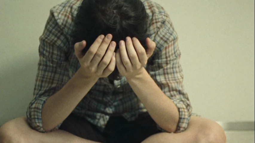 Wendy and Lucy (2008) movie still: medium — A person sits on the floor against a plain wall in an interior space. The individual hides their face…