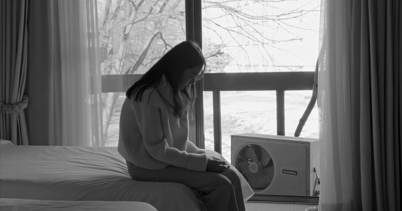 Hotel By The River movie still: medium — A hotel room with a large window looking out onto bare trees in winter. A young woman sits on the edge of a…