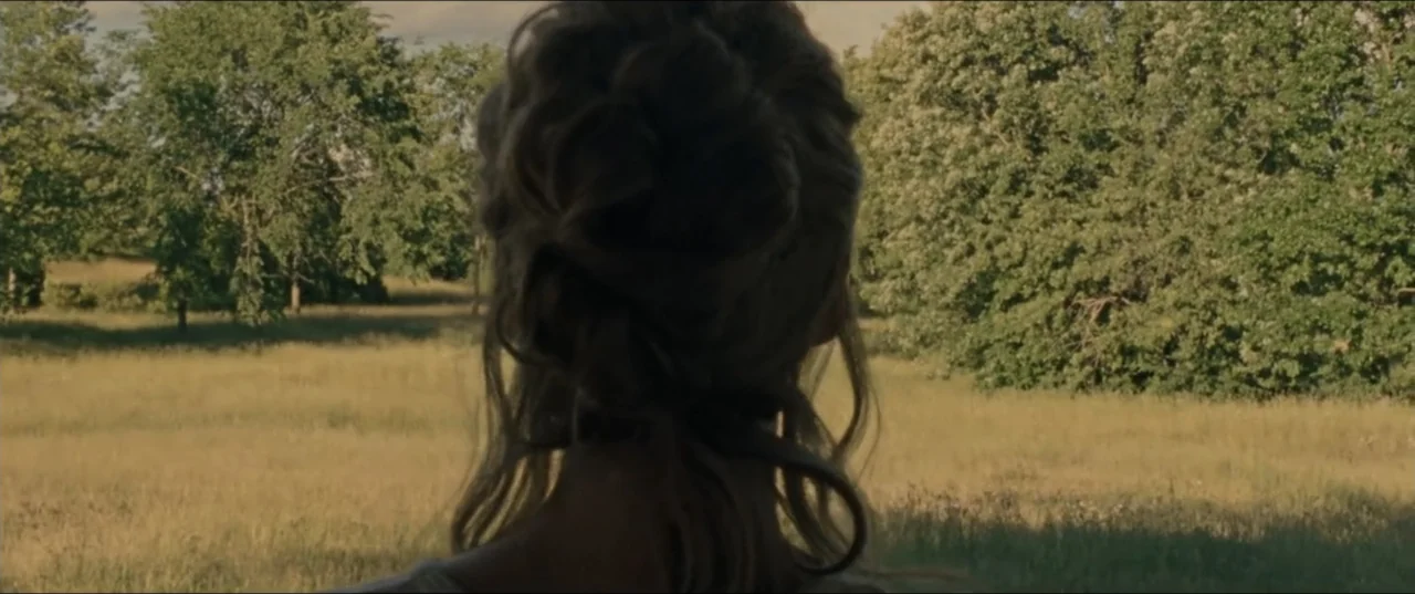 mother! (2017) movie still: medium — A grassy meadow with scattered trees stretches toward a treeline in the background. A young woman stands with…