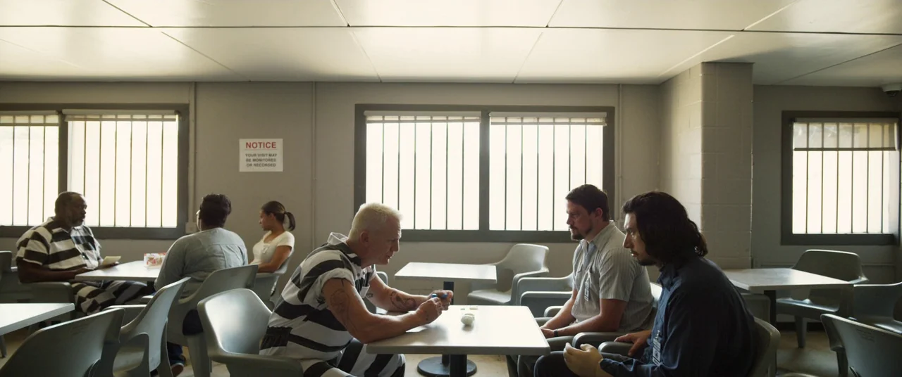 Logan Lucky (2017) movie still: wide — A prison visitation room with light grey walls, white tables, and barred windows. A group of people sit at…