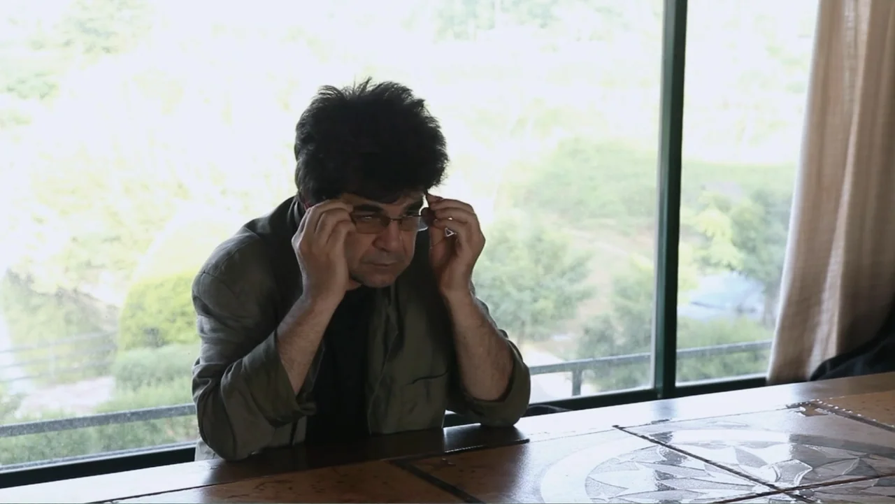 Closed Curtain (2013) movie still: medium — A man sits at a table in a sunlit interior overlooking trees. The man adjusts his glasses with both…