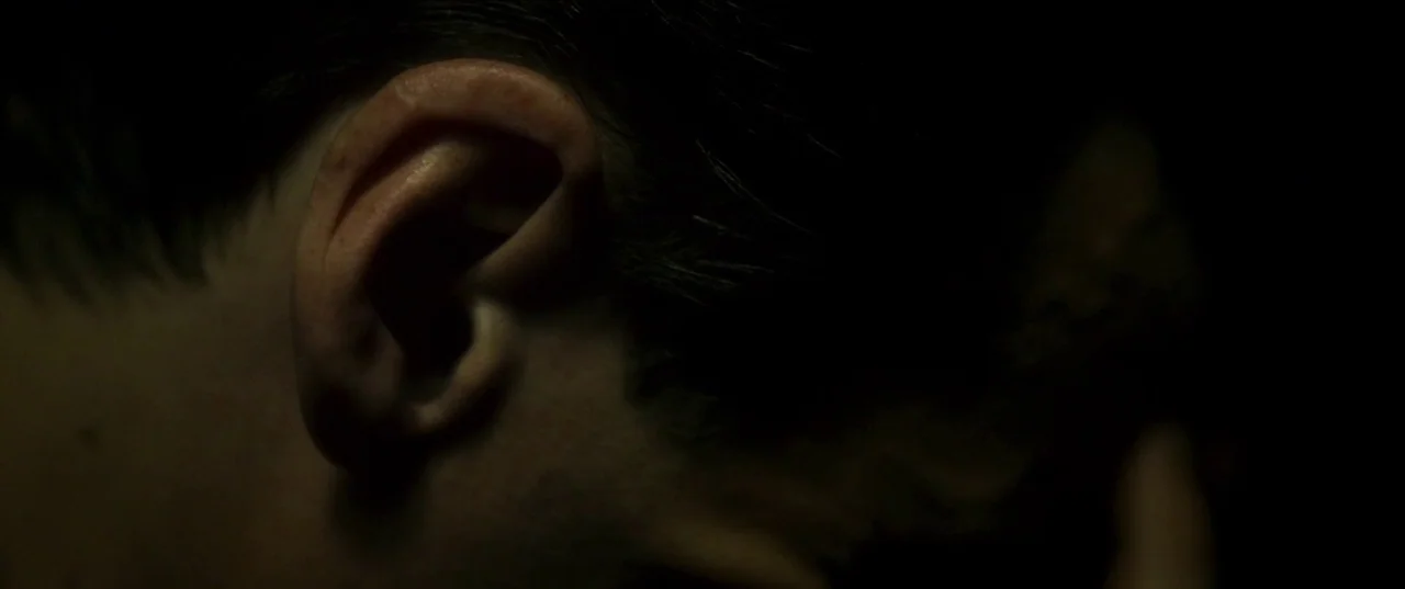 Earwig (2022) movie still: closeup — A very dark, nearly black environment showing only the side of a person's head. The focus is specifically on…