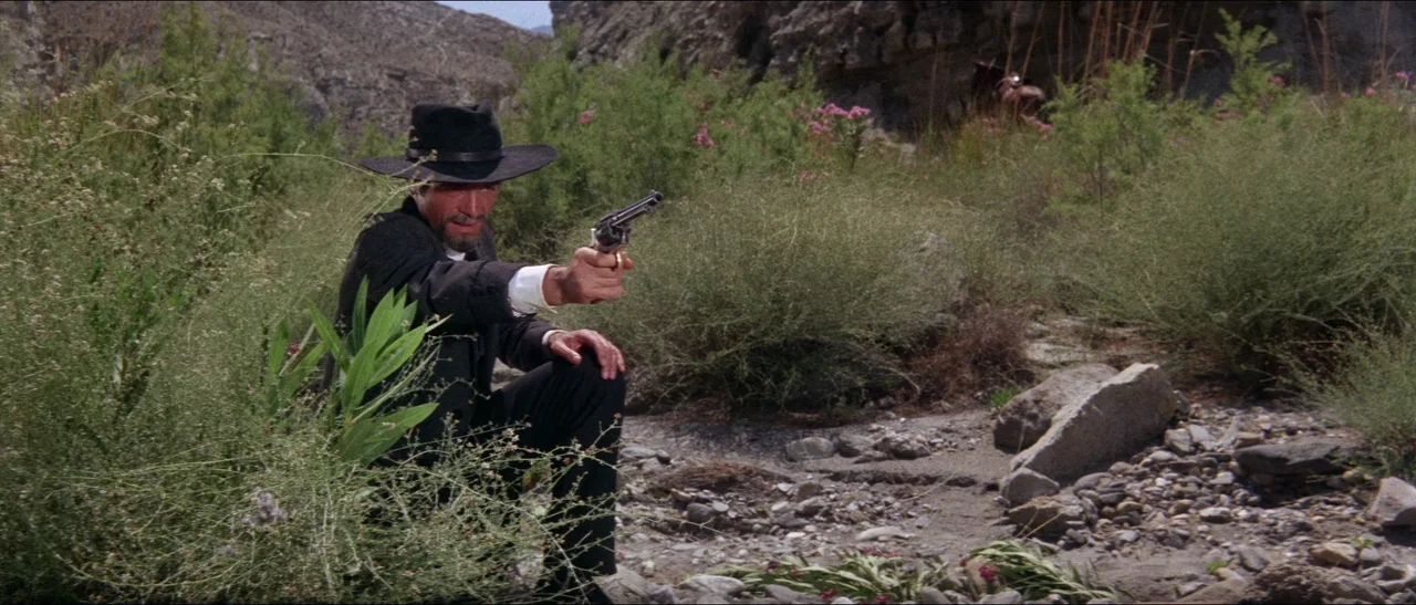 Texas, Adios (1966) movie still: medium — A man kneels within a sparse area of green bushes and desert stone. He holds a revolver aimed directly at…