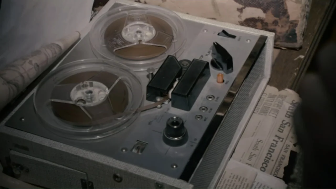 Quarantine (2008) movie still: closeup — A close-up of a vintage reel-to-reel tape recorder resting on a cluttered desk surface. Old newspaper…