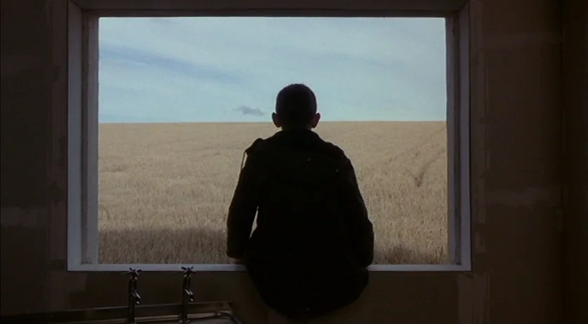 Ratcatcher (1999) movie still: medium — A person sits on the sill of a large rectangular window within an unfinished structure. Beyond the frame, an…