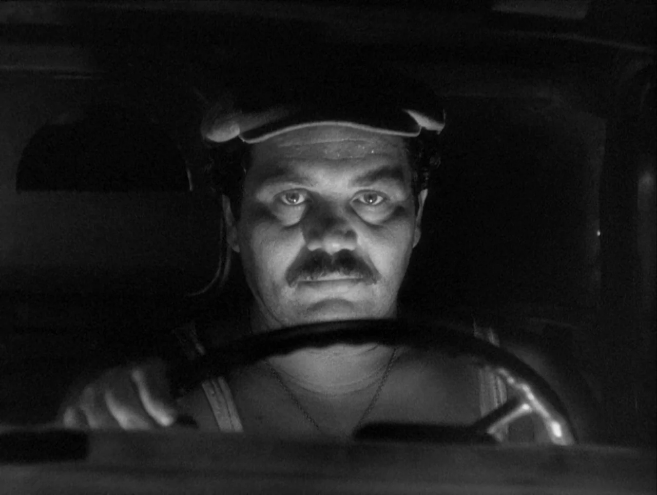 The Wages of Fear (1953) movie still: closeup — The interior of a vehicle at night is engulfed in darkness. A man wearing a flat cap holds the…