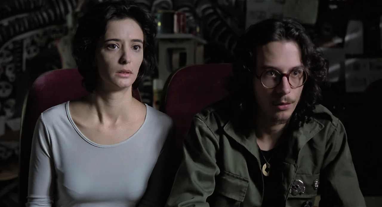 Thesis (1996) movie still: medium — Two individuals sit together in a dark room with blurred patterned walls behind them. A woman with dark hair and…