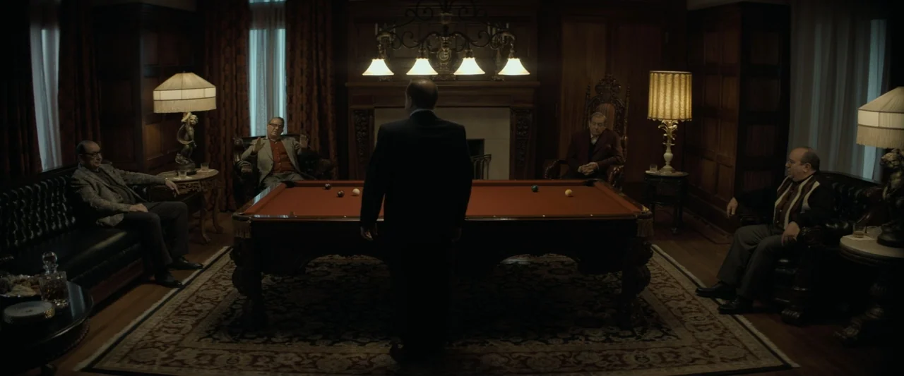 The Alto Knights (2025) movie still: wide — A dark wood-paneled room contains a large billiard table centered on a patterned area rug. Four men sit…